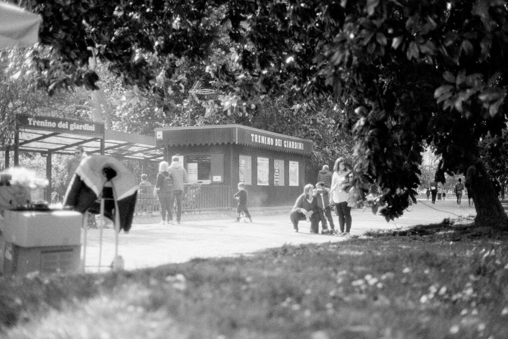 Black and white photo of a lady whispering in a child's ear with another child on a scooter in Parco Giardini Indro Montanelli, Milan, Italy